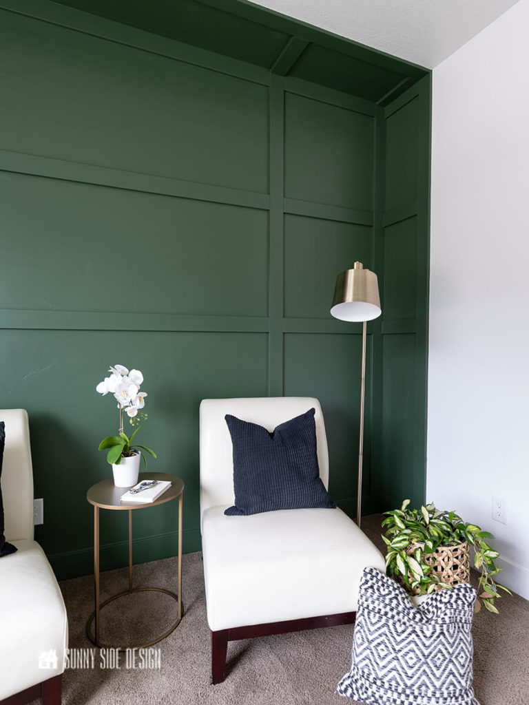 Forest green board and batten wall with the illusion of a recessed wall, white leather slipper chairs, brass lamp, brass round metal table, plants and pillows.