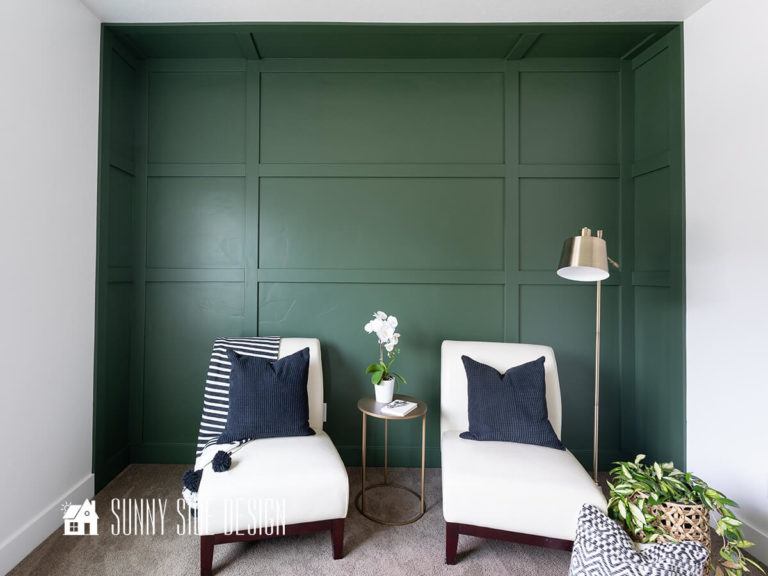Forest green DIY board and batten wall with the illusion of a recessed wall, white leather slipper chairs, brass lamp, brass round metal table, plants and pillows.