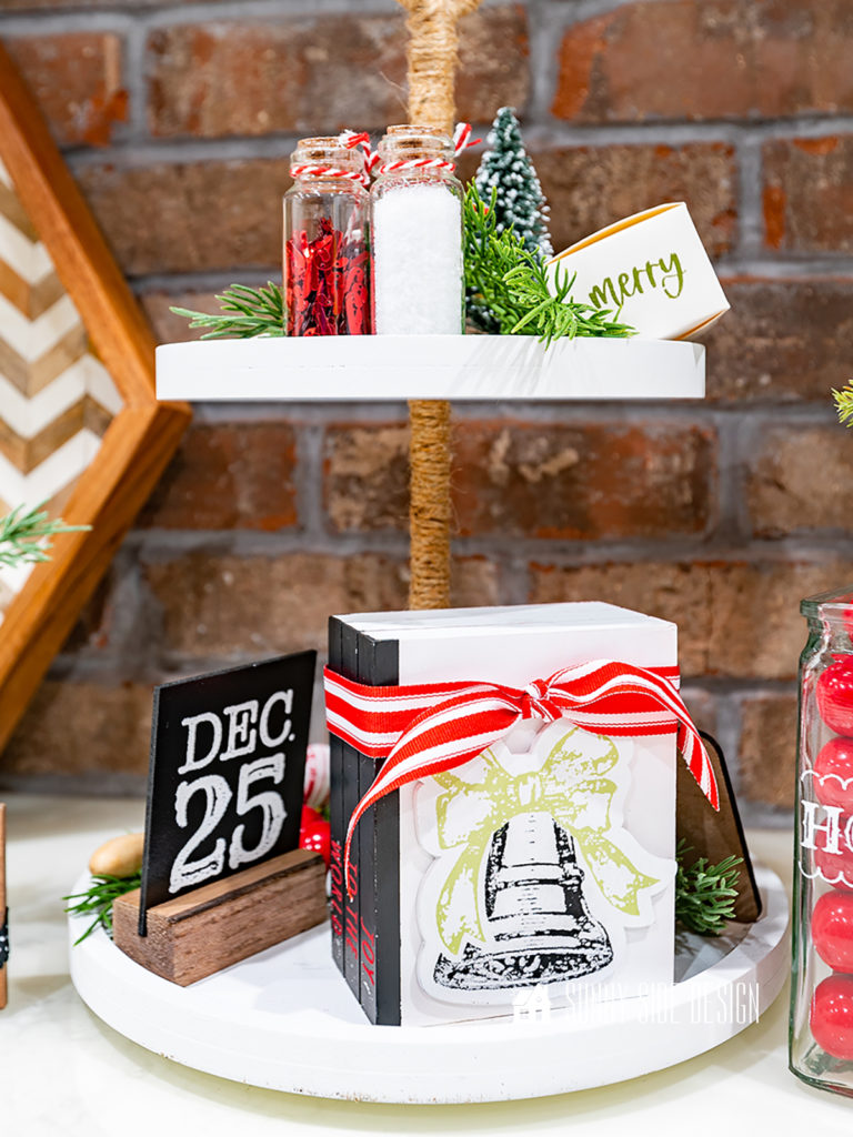 Tiered tray decor, including a book stack, chalkboard sign, greenery, bottle brush tree and small bottles filled with Christmas confetti on a white tiered tray with a jute wrapped pole.