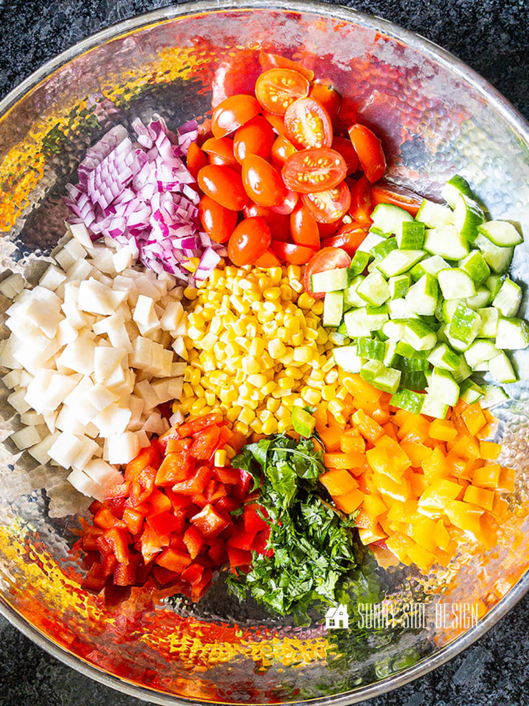 Ingredients for an easy corn salad recipe, diced jicama, red bell pepper, orange bell pepper, cucumber, red onion, halved grape tomatoes and chopped cilantro in a steel bowl.