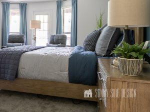 Use color schemes such as blues, grays browns to create a calling masculine interior. Modern natural wood framed bed, layered with shades of blue bedding and pillows. Accents in the room are black and gray to ground the space.