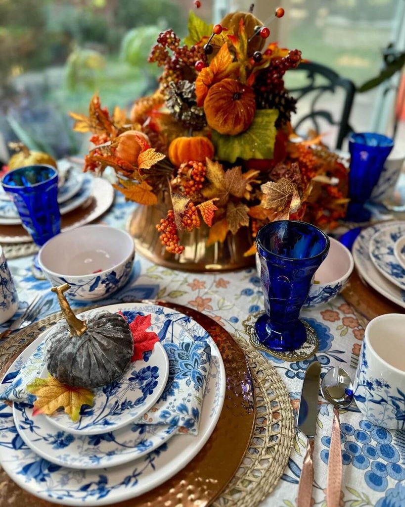 Blue and white dishes, napkins set the tone for this rich Thanksgiving table decor. Plates are topped with a blue velvet pumpkin, cobalt blue goblets and copper colored charger. The centerpieces is shades of oranges berries, leaves and pumpkins in a bronze containter.