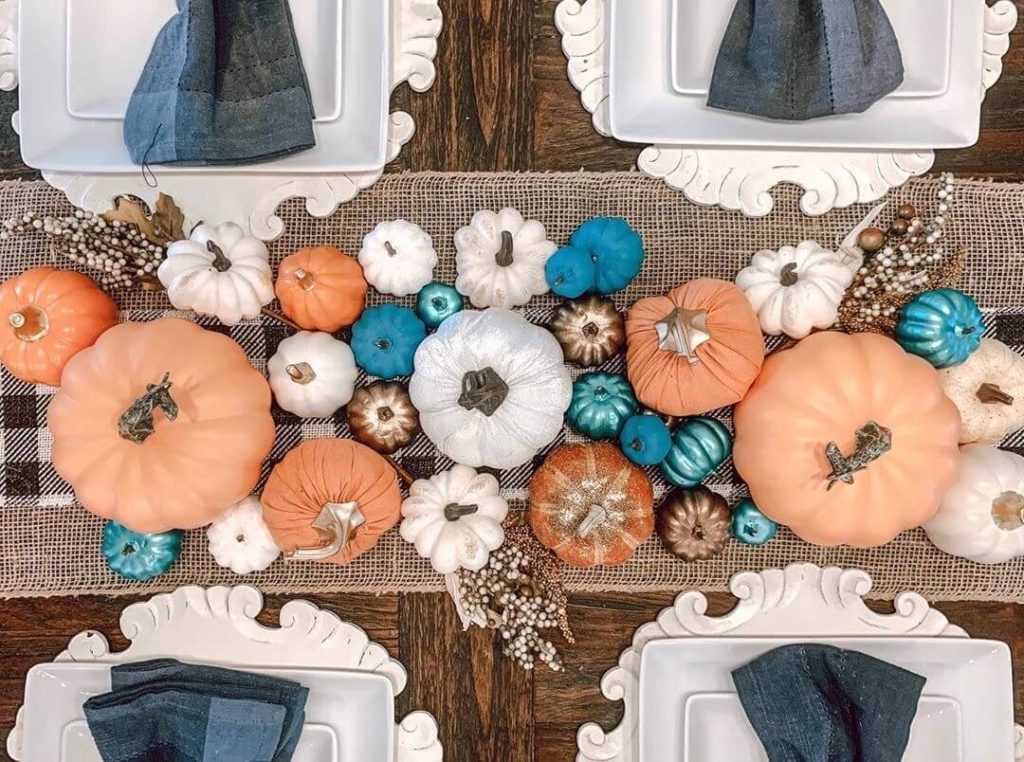 Colorful pumpkin centerpiece with a variety of orange, peach, bronze, white, hold and teal pumpkins on a burlap table runner. Each place setting had white plates on a white wood charger with a chambray napkin.