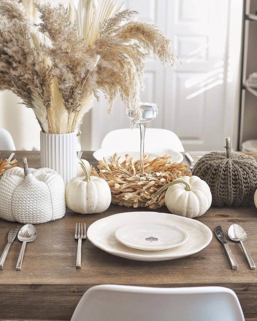 Boho inspired Thanksgiving tablescape with white vases filled with pampas grass, pumpkins and corn husk wreaths also form the centerpiece. Each place setting features white places on a rustic wood table.