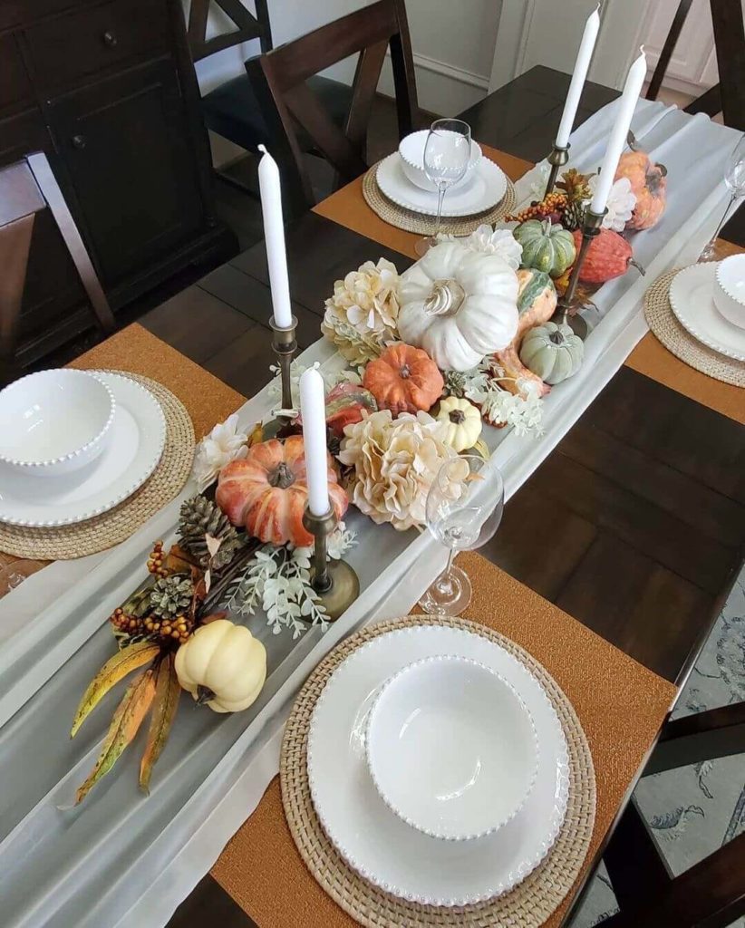 Rich fall colored tablescape featuring a pumpkin and floral centerpiece that lies low on the table with vintage brass candlestick with tall white tapers. Each place setting features white places on a wicker charger and a rust colored placemat.