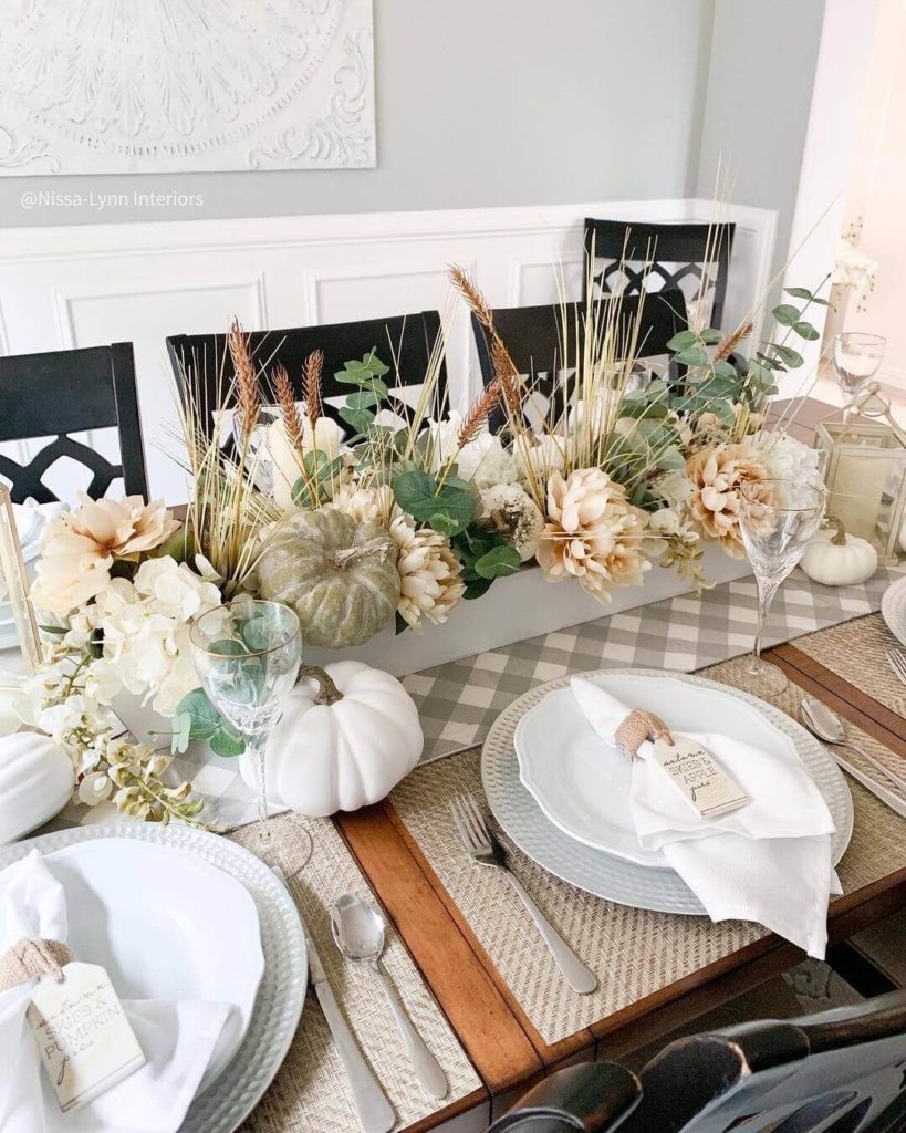 Nature inspired Thanksgiving table decor with natural woven placemats with a textured charger and a white plate, topped with a white napkin with a jute napkin ring with an attached fall tag. The centerpiece is made of assorted neutral pumpkins, florals and grassed in a white wooden box.