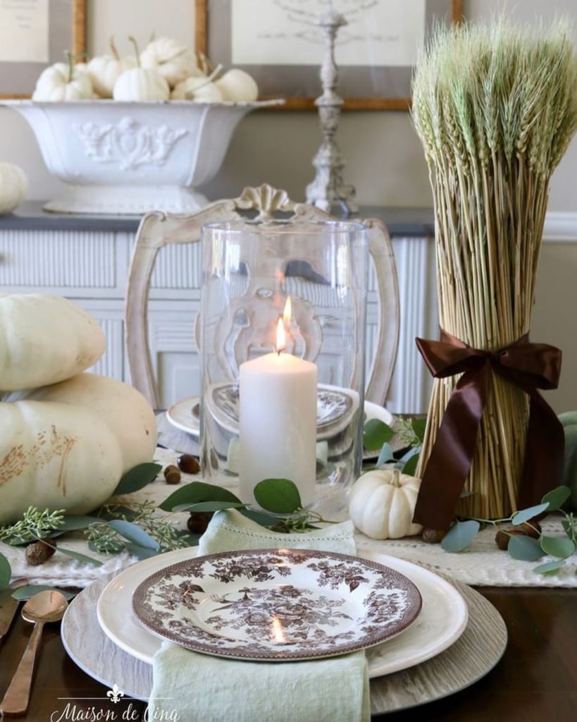Neutral Thanksgiving table decor, wheat stalks tied with a brown satin ribbon, white pillar candle in hurricane, white stacked pumpkins, and eucalyptus form the centerpiece. The place setting features a white plate on a wood grain charger, topped with a sage green napkin and a brown transferware plate.