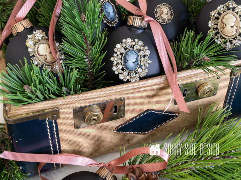 Easy Christmas Ornament to Make, black round ornament with a rhinestone cameo, antique gold ornament cap with a dusty rose ribbon. Styled in a vintage train case with fresh evergreens.