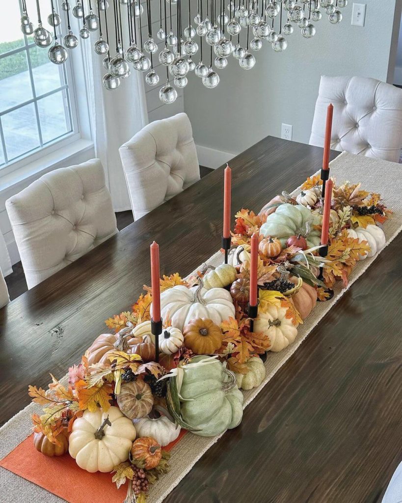 A festive fall centerpiece on a natural linen table runner topped with an orange table runner and then a variety of pumpkins in hues of white, tan, brown, green and orange mixed with oak leaves and berries. Black candlesticks with orange taper candles.