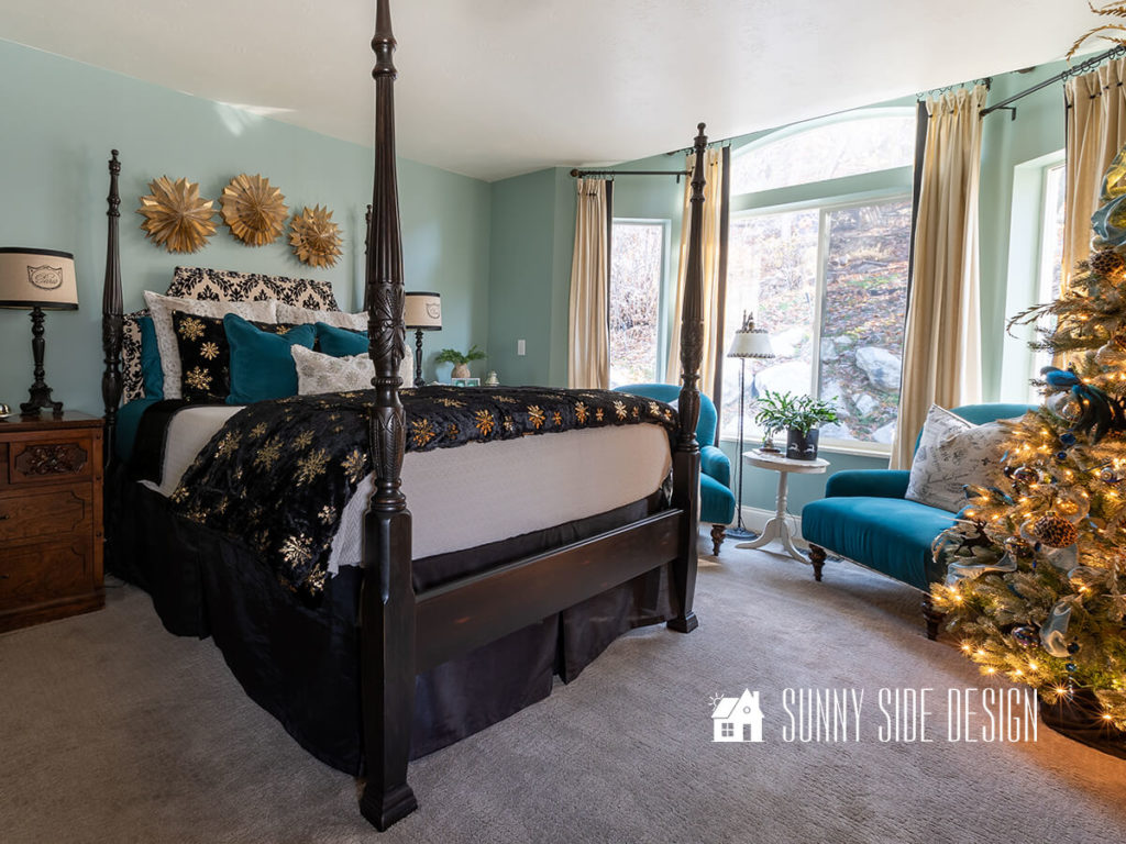 Bedroom is decorated for Christmas with a black minky and gold snowflake duvet with additional snowflake pillows on the bed. Above the bed 3D gold snowflakes are hung and on the night stand are a variety of glass Christmas trees. A full sized flocked Christmas tree is decorated in gold, black and peacock blue. Blue velvet chairs are placed in the bay window.