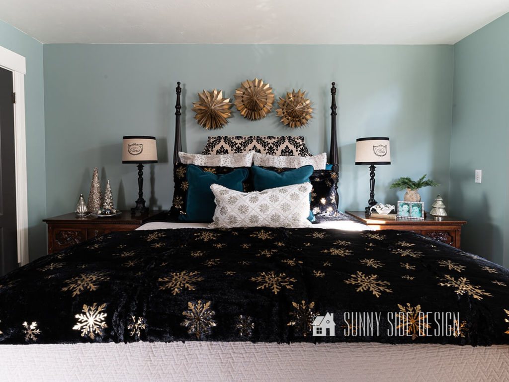 Bedroom is decorated for Christmas with a black minky and gold snowflake duvet with additional snowflake pillows on the bed. Above the bed 3D gold snowflakes are hung and on the night stand are a variety of glass Christmas trees.
