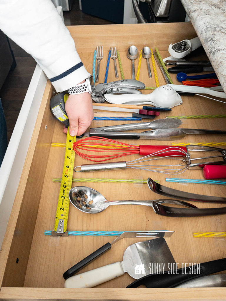 Various kitchen utensils in drawer, using straws as divider to plan out how to organize drawer. Woman's hand with tape measure finds distance for section.