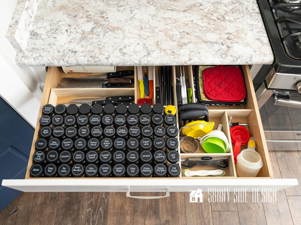Custom wood DIY drawer dividers in kitchen spices alphabetized, knives, measuring spoons and cups, funnels, slicer, scissors, hot pads neatly organized in kitchen drawer.