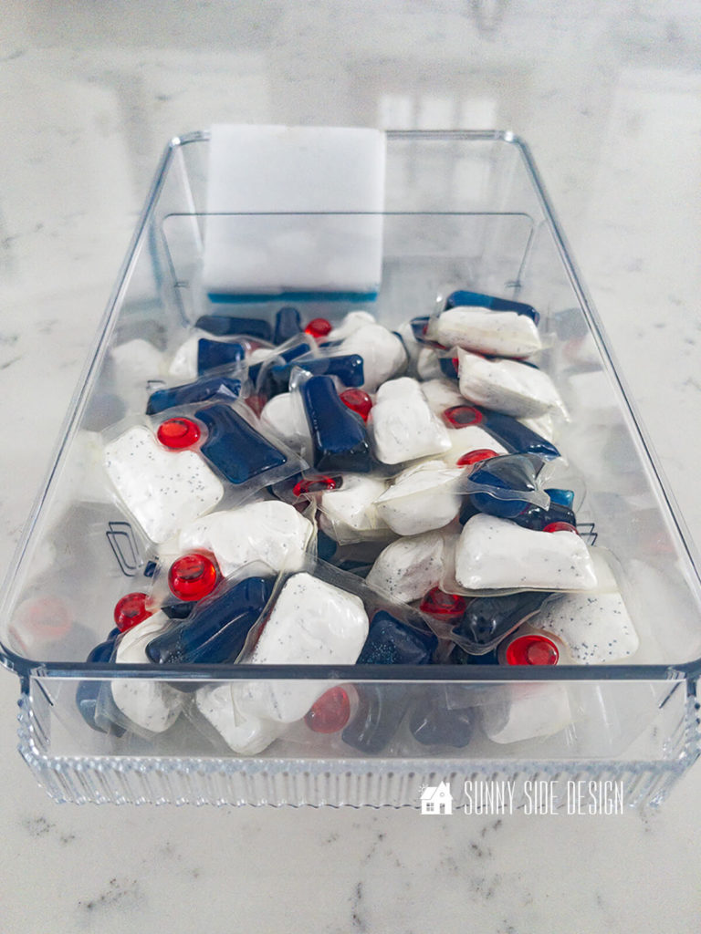 Dishwasher cleaning tabs are stored in a clear acrylic bin under the kitchen sink.