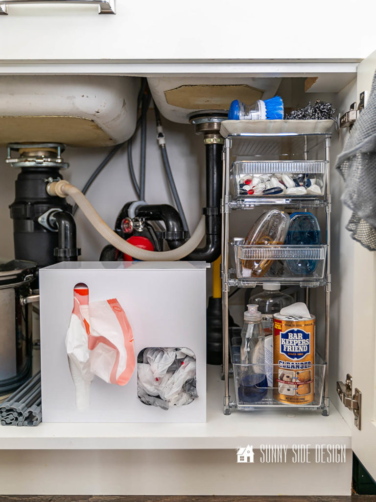 The best tips to organize under the kitchen sink: tiered storage rack with sliding bins holds kitchen cleaning supplies, white plastic contain for storing trash and grocery bags.