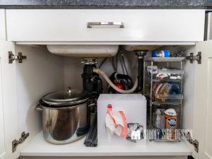 The best tips to organize under the kitchen sink: tiered storage rack with sliding bins holds kitchen cleaning supplies, white plastic contain for storing trash and grocery bags, drying rack, and stock pots.