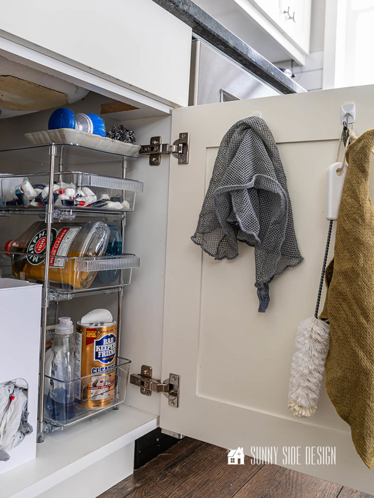 The best tips to organize under the kitchen sink, command hooks on cabinet door for cleaning cloths and bottle brush, tired storage trays for cleaning supplies.