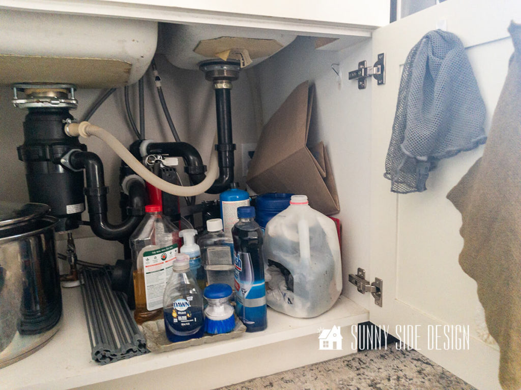Under kitchen sink before organization. Random clean supplies, paper bags, old milk jug for grocery bag storage, pans and drying rack.