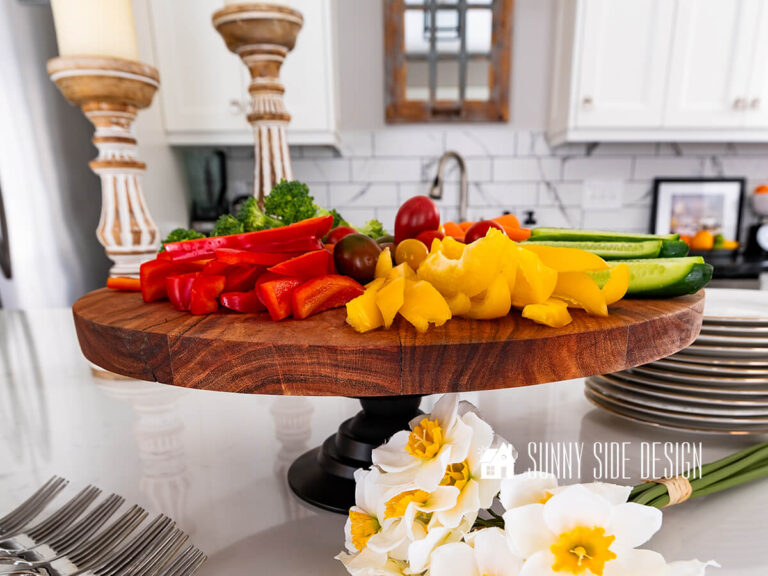 Styled pedestal serving tray on kitchen island with wood candlesticks, daffodil bunch with assorted vegetables on the pedestal serving tray.