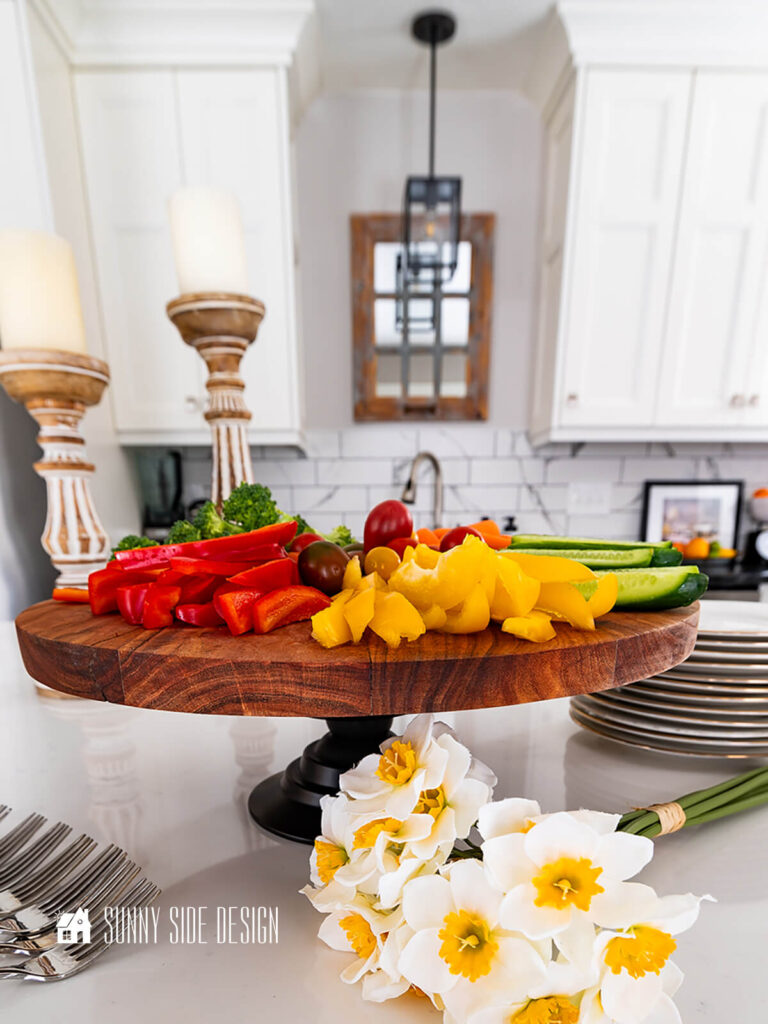 Styled pedestal serving tray on kitchen island with wood candlesticks, daffodil bunch with assorted vegetables on the pedestal serving tray.