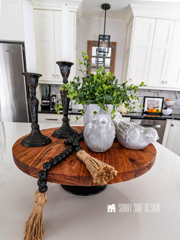 Styled pedestal serving tray on kitchen island with concrete birds, potted plant, black candlesticks and a black bead garland.