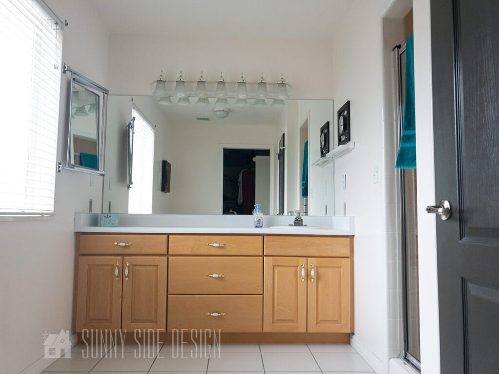 Master bathroom when home was purchased, white tile floor with green grout, large maple vanity with single sink on right, white laminate countertop, large plate glass mirror, white vanity light, shower behind wall on right.