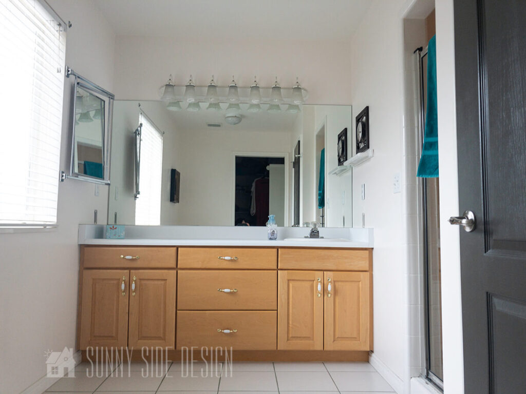 Master bathroom when home was purchased, white tile floor with green grout, large maple vanity with single sink on right, white laminate countertop, large plate glass mirror, white vanity light, shower behind wall on right.