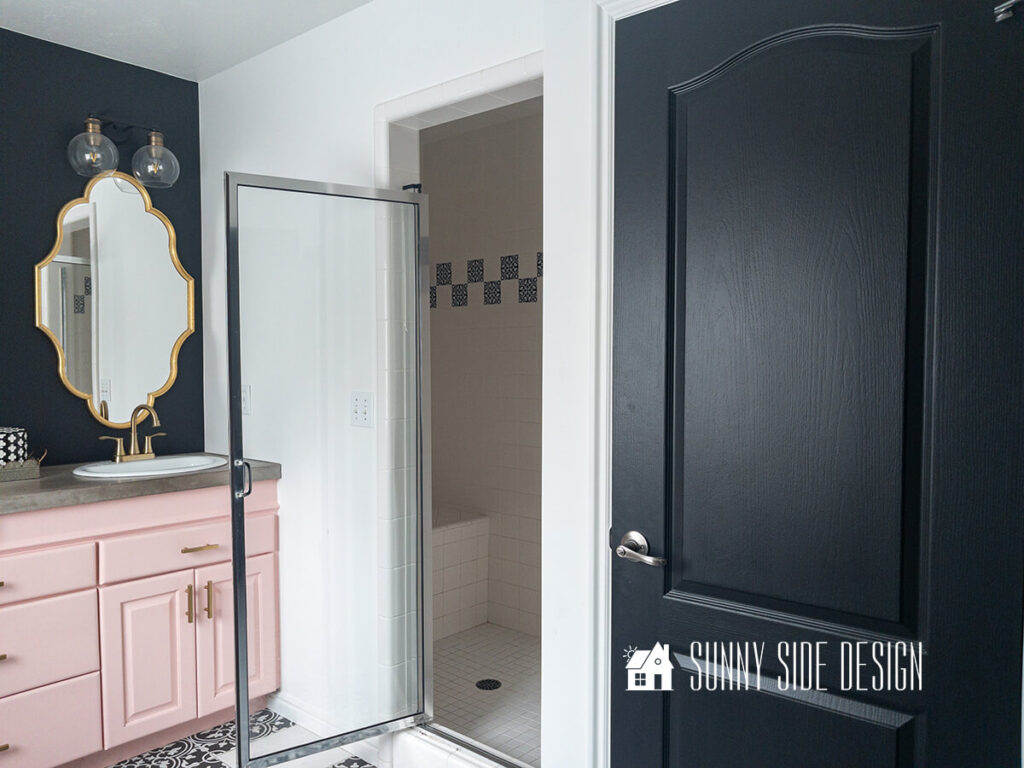 Cosmetic updates to Master bathroom, with black and white vinyl decals on floor, white paint, black door, pink painted vanity, gold hardware, mirrors and lights, faux fur ottoman, black accent wall above vanity. Walled off shower with vinyl decals covering pink and green tile.