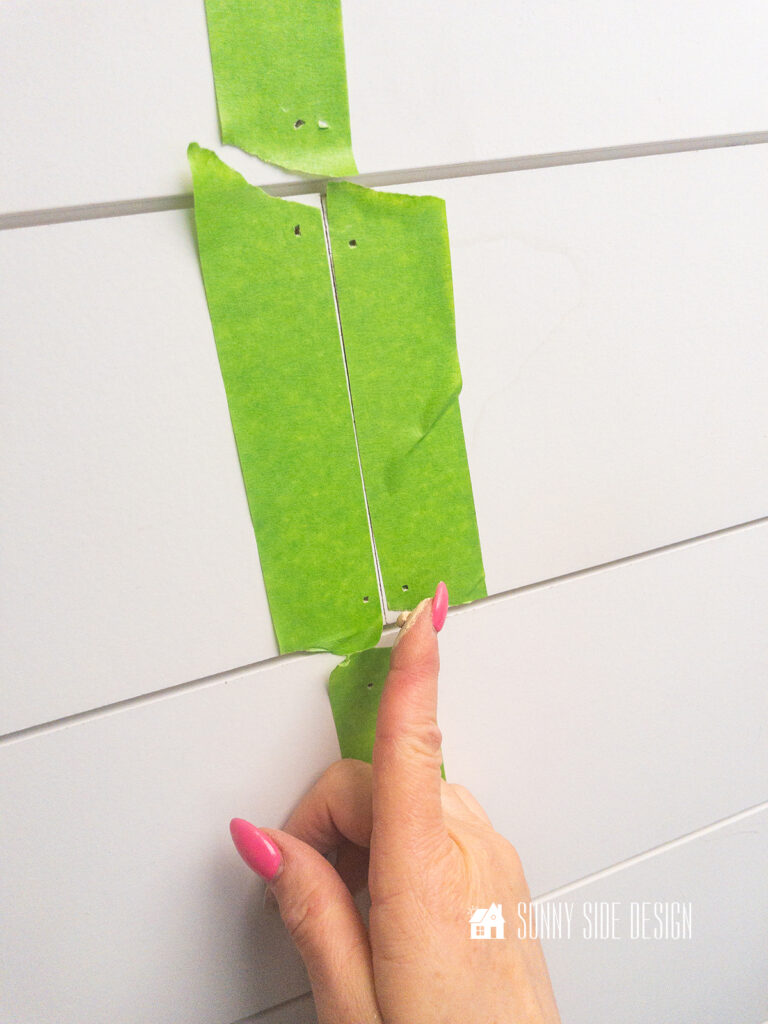 Woman's finger applies wood filler through the hole in the painters tape on the shiplap ceiing.