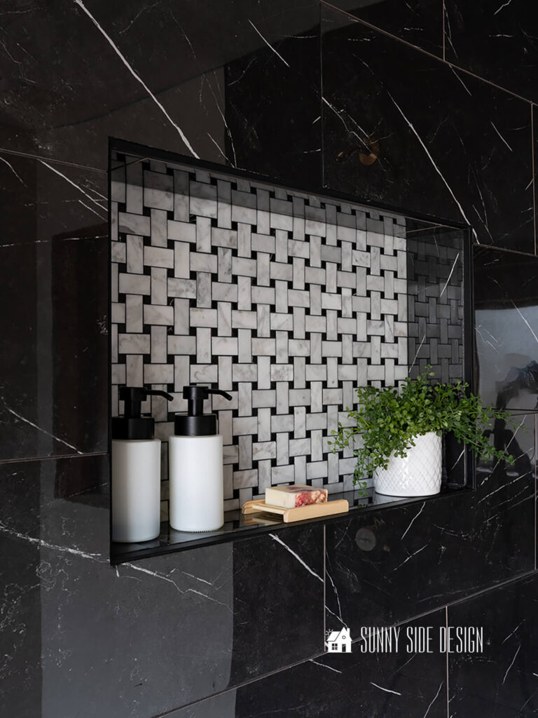 Tiled shower niche with Carrara marble basket weave tile and black marble shower wall tile. Styled with pink marble soap, black and white pump bottles, and a plant.