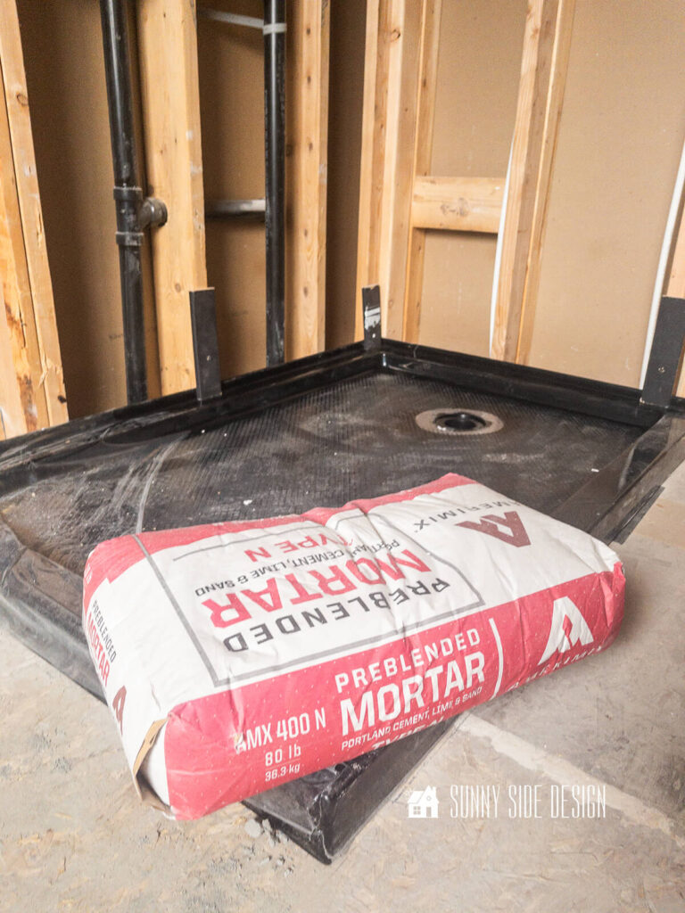 Bag of mortar is used to weigh down the black prefab shower pan while mortar dries in this bathroom remodel.