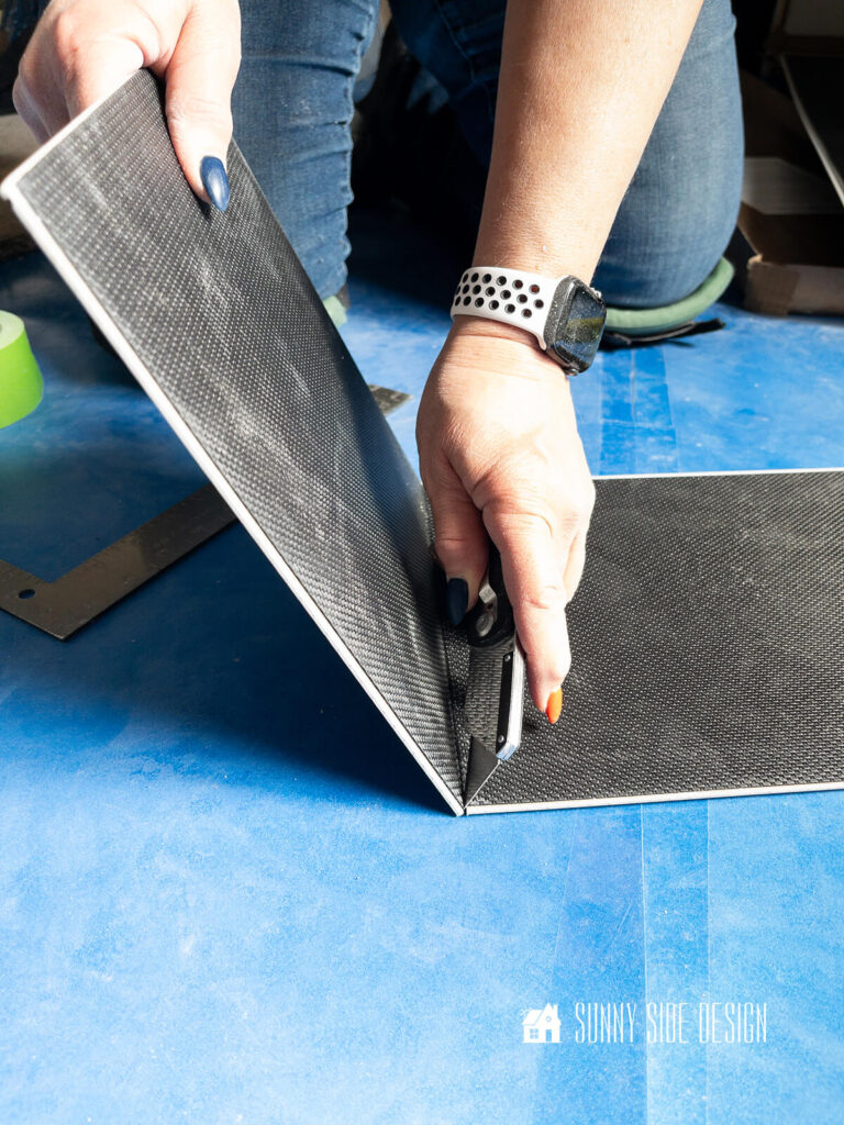 After vinyl plank flooring is scored and snapped, woman cutes through back padding with a utility knife.