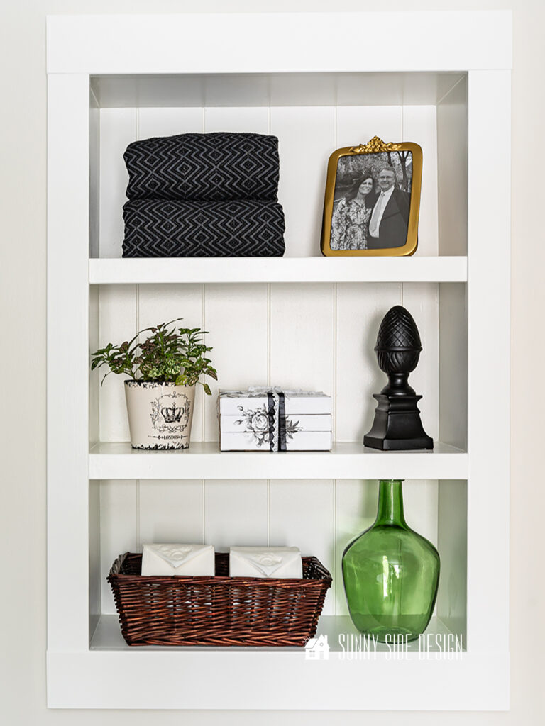 Installed and decorated recessed shelf in a wall with black towel, gold photo frame, potted plant, book stack, black finial, green glass vase, basket with extra toilet paper.