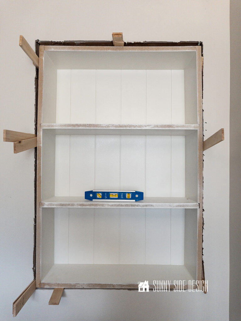 DIY shelf placed in wall with shims placed on the sides to level up the shelf before it is secured to the studs with nails.