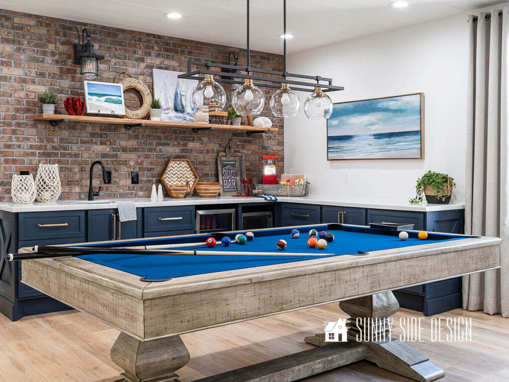 Pool table with lights and wet bat with navy blue cabinets and brick wall. Assorted coastal decor.