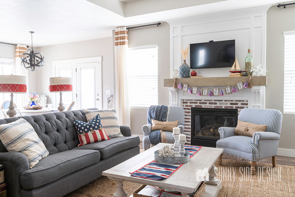 Living room with a craftsman style fireplace and beam mantle and red brick surround, blue and white striped chairs, white coffee fable, sisal rug, grey sofa.
