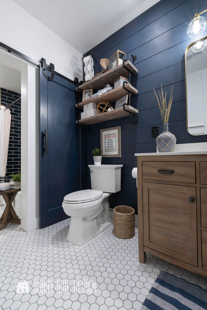 Home tour modern coastal bathroom with a navy blue shiplap wall, white hexagon floor, reclaimed wood vanity with white quartz countertop. Rustic shelves with wood and metal are hung over the toiled and decorated with coastal decor.