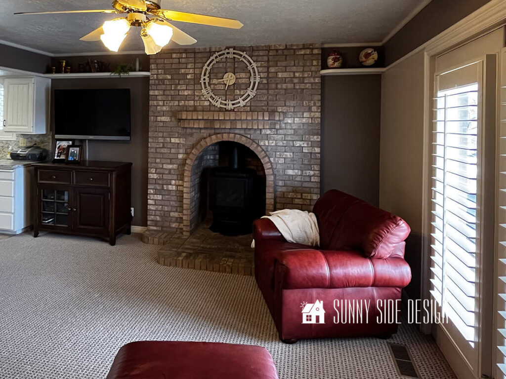 Dark and dated room before makeover. Dark oversized mahogany colored chair pushed against dark brown and beige walls. Brick fireplace with wood burning stove, dark wood media cabinet and TV. The only light is a ceiling fan light.