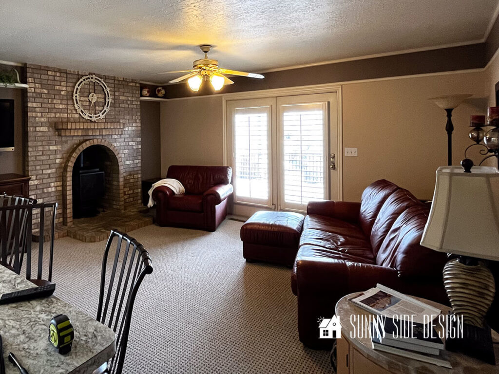 Dark and dated room before makeover. Dark oversized mahogany colored chair pushed against dark brown and beige walls. Large over-stuffed mahogany sofa and ottoman. Brick fireplace with wood burning stove. The only light is a ceiling fan light.