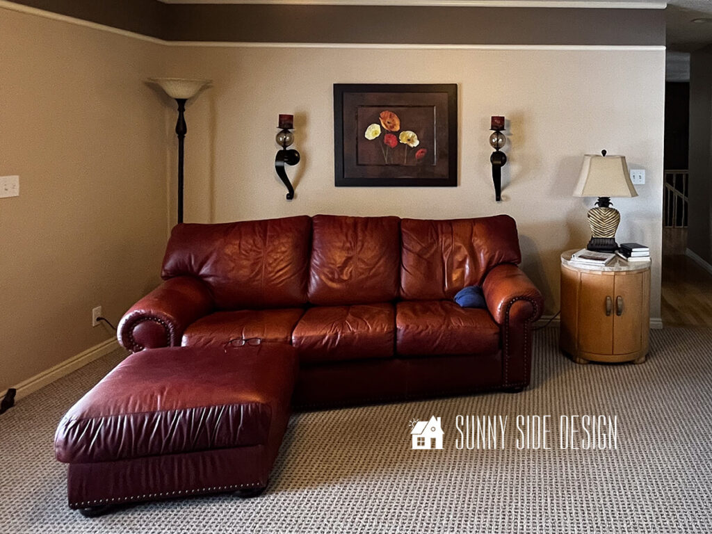 Dark and dated room before makeover. Large over-stuffed mahogany leather sofa and ottoman. Angled in room with beige walls and a dark brown painted boarder with white trim at the ceiling.