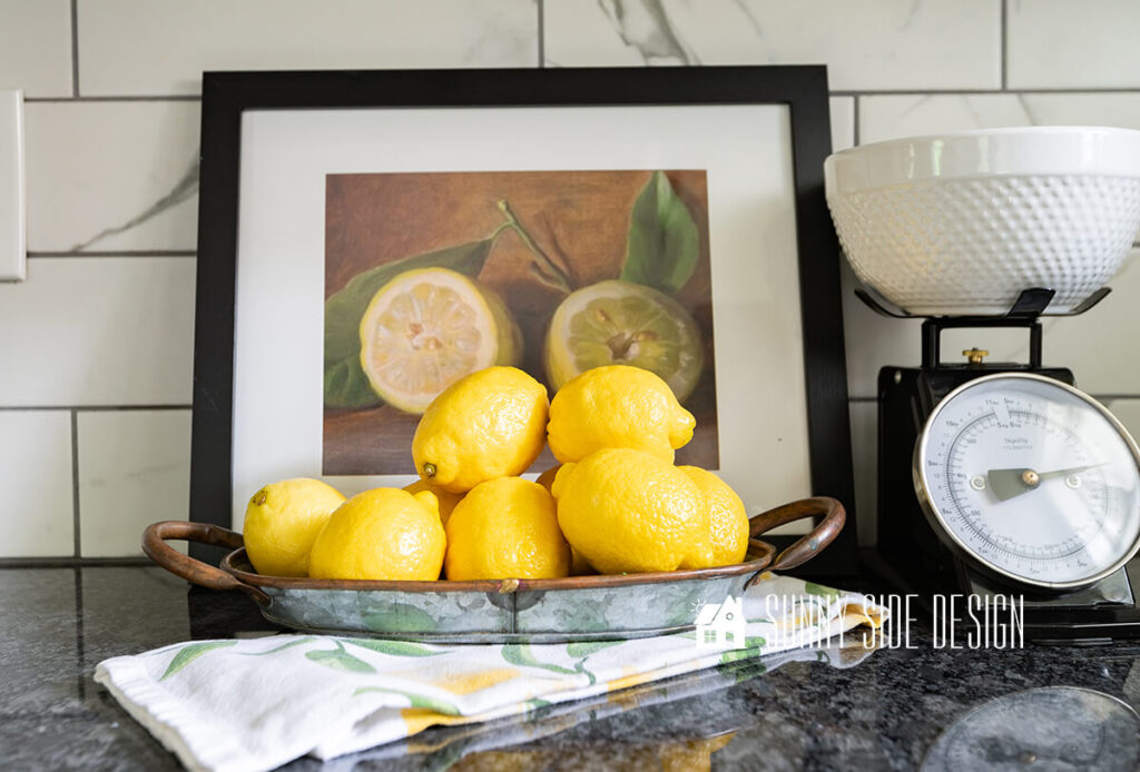 Free printable wall art in a modern black frame with a white mat. Styled in a kitchen with a tray of lemons on a lemon towel.