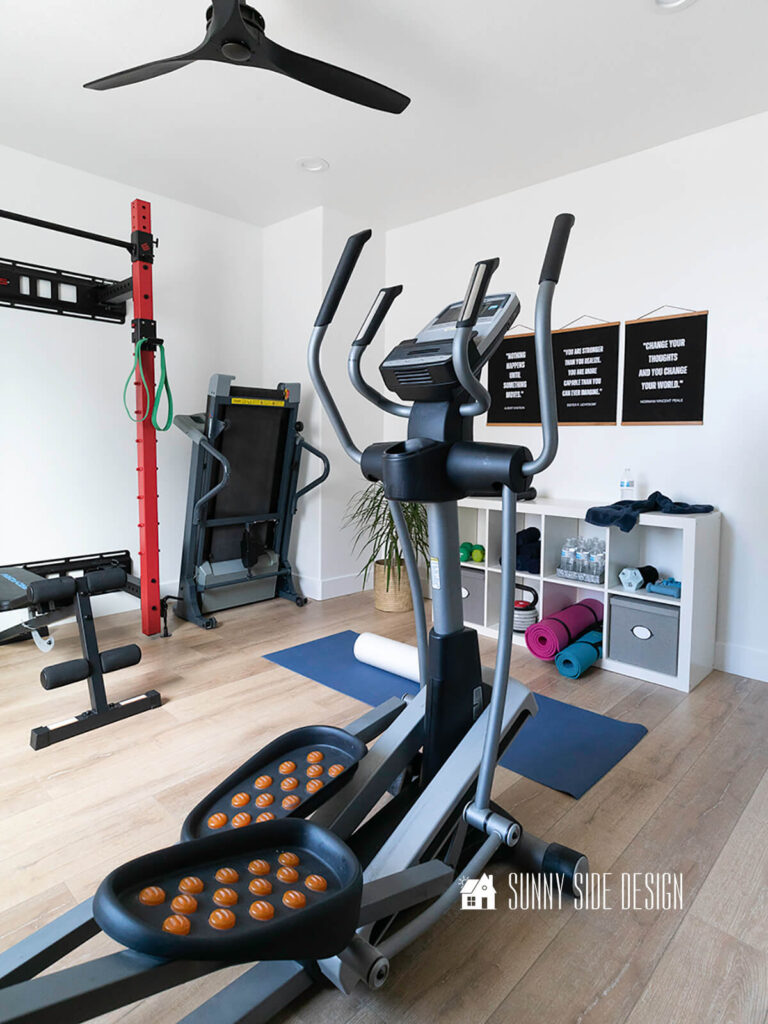Home gym with motivational quotes framed on the wall, white oak floor, storage cubbies for weights, towels and other workout equipment. Yoga mat, elliptical, tread mill.