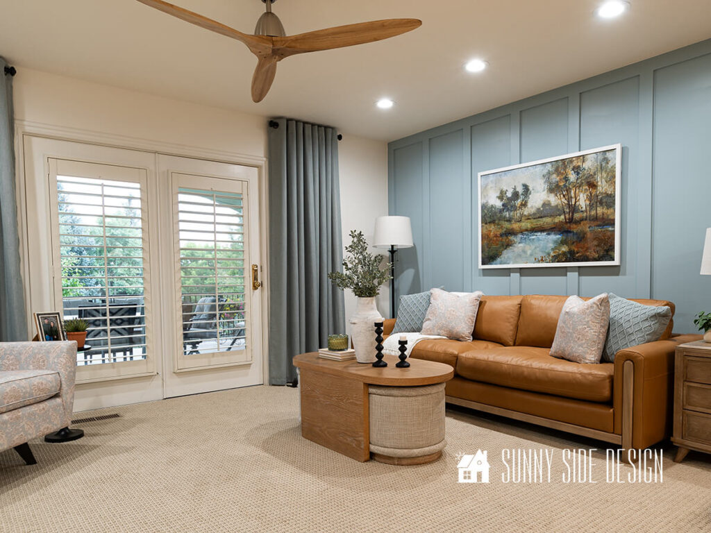 Walls, ceiling and trim moulding are painted pure white and a light blue accent wall with board and batten adds contrast and warmth to the room. Caramel colored leather sofa with oak wood trim, oak coffee table with tan ottomans, colorful art hung above the sofa.