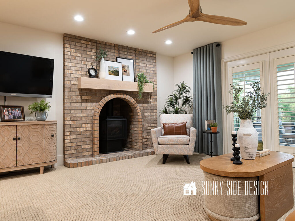 Six can lights are installed in the room with adjustable kelvin color temperature to make the light brighter and whiter. View of brick fireplace with French oak mantle and blue and beige paisly side chair. Mantle decorated with white framed art, plants, clock.