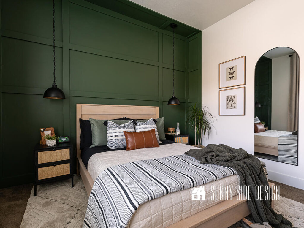 Bedroom home tour, green board and batten wall that extends to the side walls, white oak bed frame with white, black and green bedding. Black pendant lights are hung from the ceiling over the black and rattan nightstands. Framed art and a black framed arched mirror.