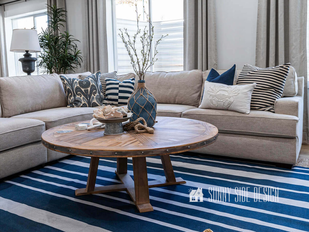 An old wood kitchen table is upcycled into a large round coffee table in this family room. Large tan sectional with a navy blue and white striped rug.