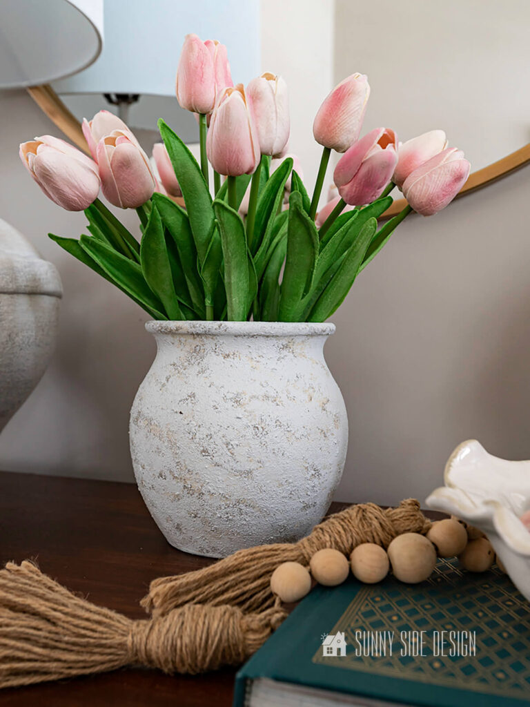 Thrift store canister is transformed with paint and salt wash, creating a textured vase. Vase styled with pink tulips and a natural wood bead garland.