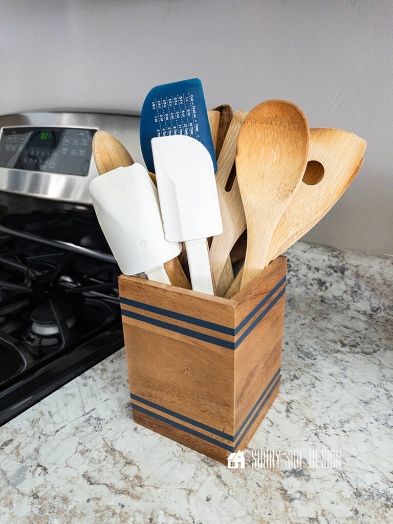 Thrifted wood vase is upcycled into a kitchen tool caddy. Imperfections were sanded out and fun navy blue stripe was painted on the wood vase.