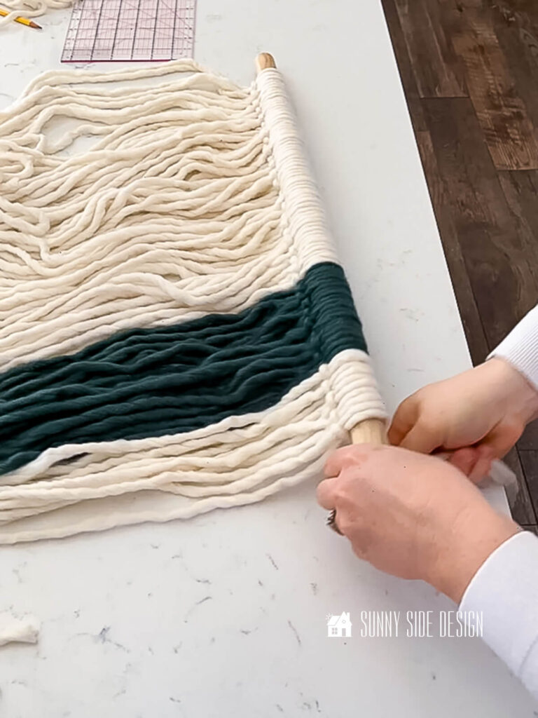 Woman loops roving yarn around a dowel to form a larks head knot for the yarn wall art.