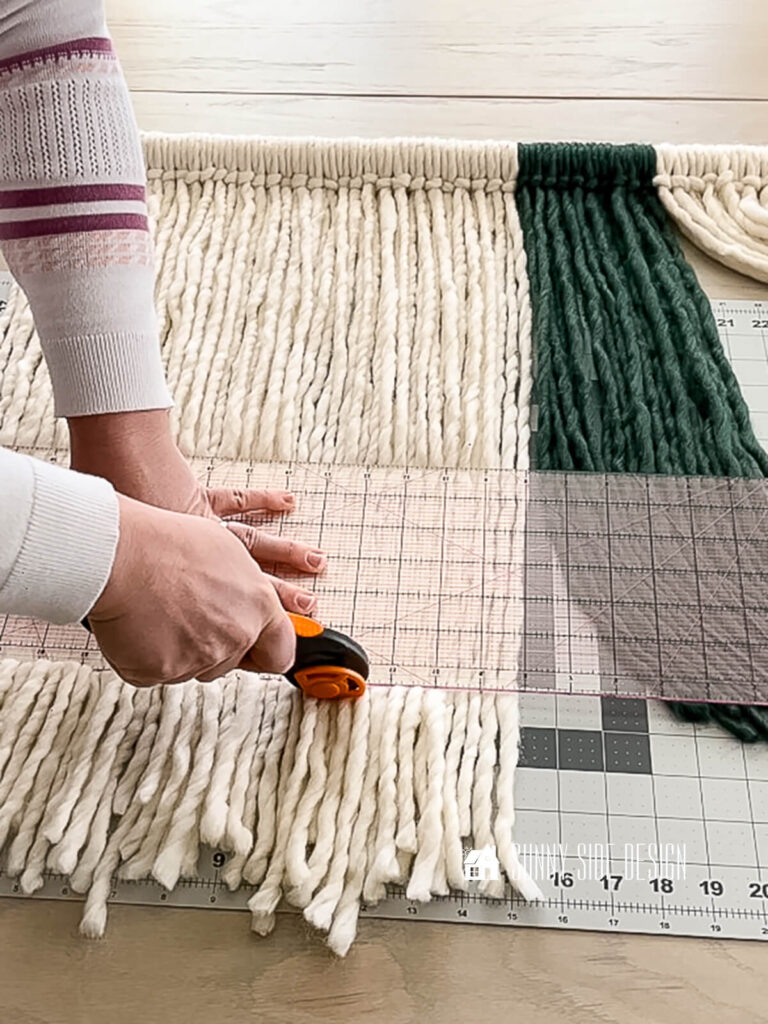 Woman uses a rotary cutter and a quilting ruler to trim the lower edge of the yarn wall art.